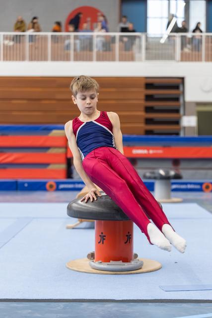 Young gymnast performs on pommel horse with focused expression, legs extended in mid-routine at indoor gymnastics venue