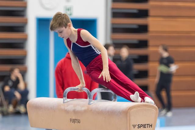 Young gymnast in burgundy and blue performs pommel horse routine with focused expression at indoor gymnastics event
