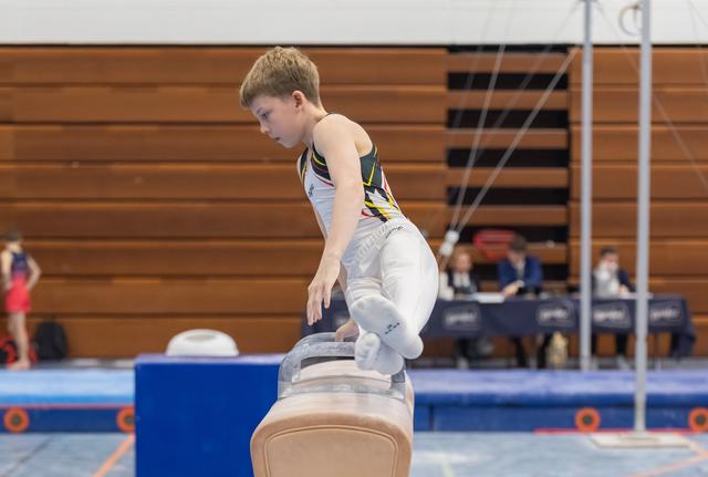 Young gymnast performing on pommel horse with focused concentration, legs positioned during routine at indoor gymnastics meet