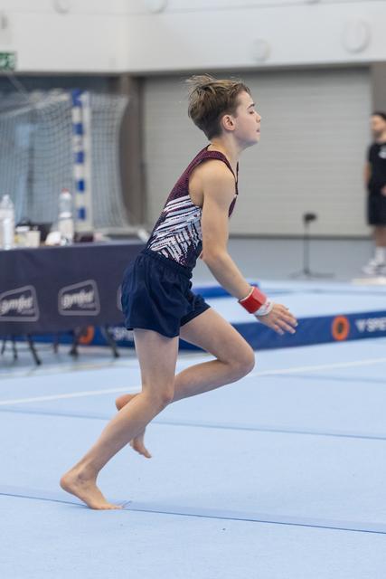 Young gymnast performing floor exercise routine barefoot on blue mat, arms extended in focused concentration