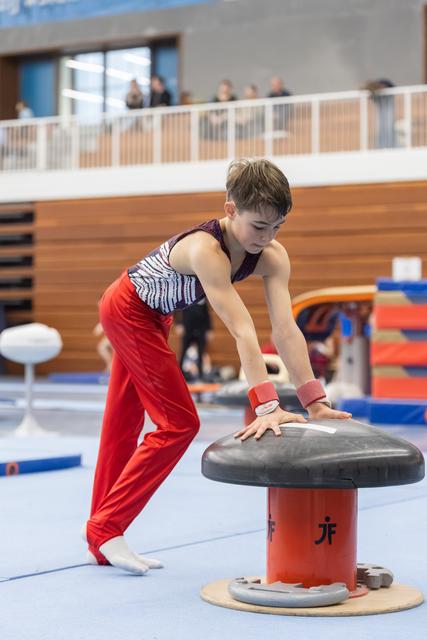 Young gymnast in red pants practices on mushroom apparatus, leaning forward with focused concentration in indoor training facility