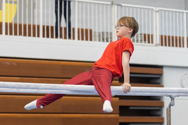 Young gymnast with glasses performing a support hold on parallel bars, focused expression, wearing red outfit in gymnasium