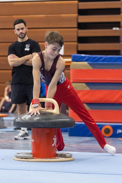 Young gymnast in burgundy leotard and red pants practices on pommel horse while coach observes in training facility