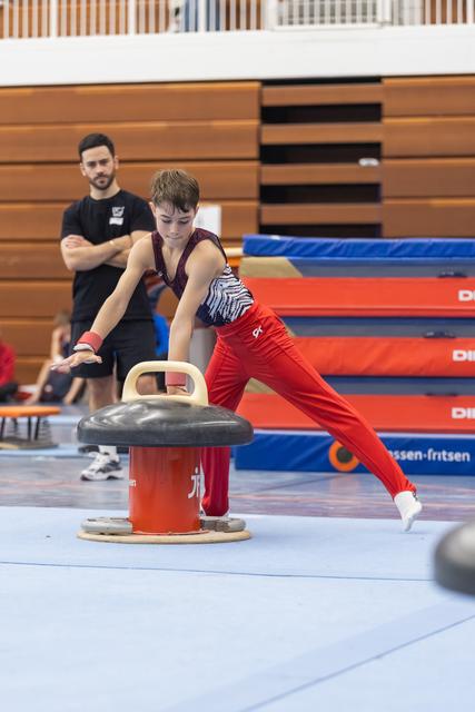 Young gymnast in red pants practices pommel horse technique while coach observes in background at training facility