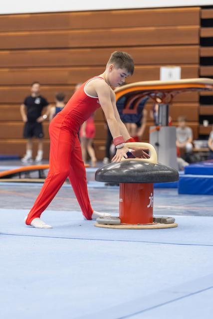 Young gymnast in red uniform practices on vault apparatus, hands positioned on pommel grips with focused concentration