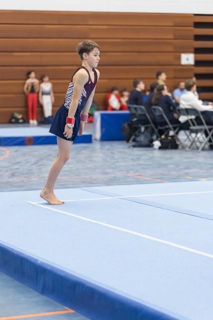 Young gymnast stands focused on floor mat, awaiting start of routine at indoor gymnastics meet