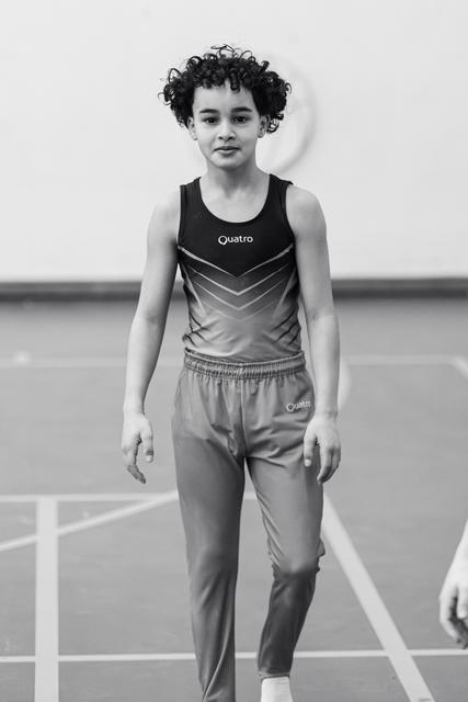 Young gymnast with curly hair stands confidently in Quatro uniform on gym floor, poised and ready for performance