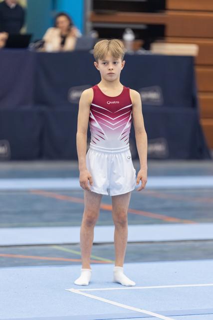 Young gymnast in burgundy and white leotard stands at attention on blue floor mat, focused and ready to perform