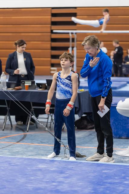 Young gymnast in blue leotard stands attentively beside coach in matching jacket, preparing for routine at gymnastics meet