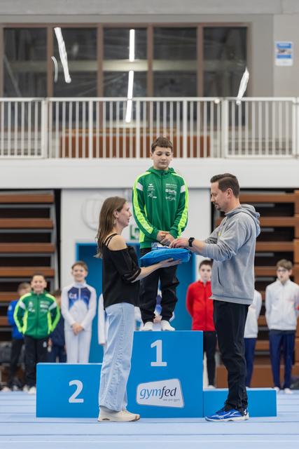 Boy in green tracksuit stands on first place podium while officials present medal at gymnastics awards ceremony