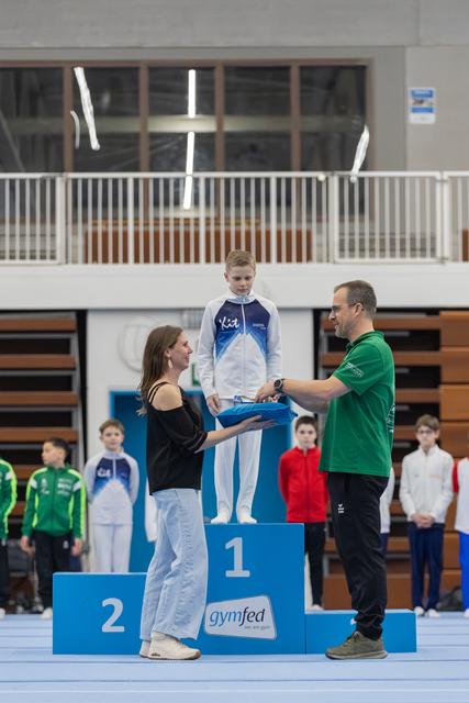 Young gymnast stands on first place podium receiving medal from coach in green shirt while woman assistant looks on