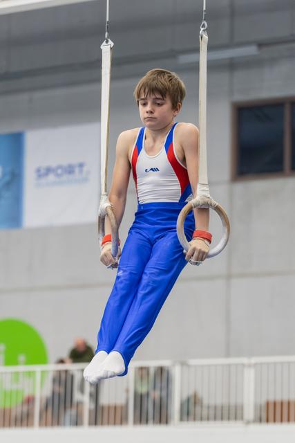 Young gymnast in blue and white uniform performing a still rings routine with focused concentration in an indoor gymnasium