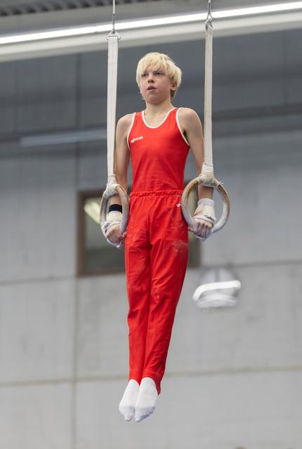 Young gymnast with blonde hair performs still rings routine in red uniform, displaying focus and control at training facility