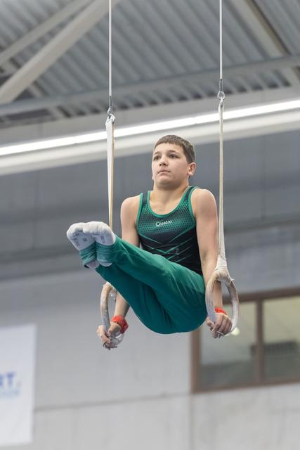 Young gymnast in green performing L-sit position on rings, focused expression, training facility with industrial ceiling