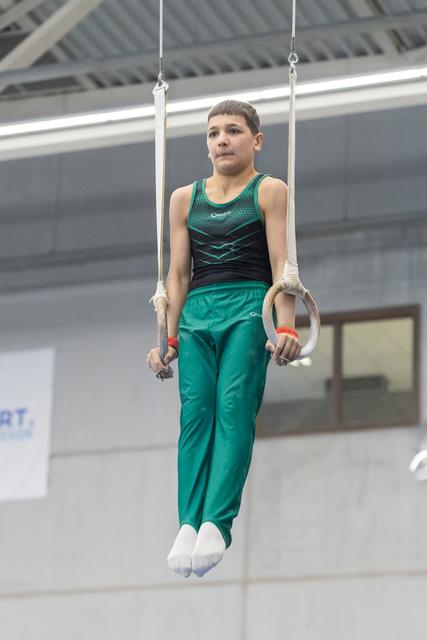 Young gymnast in teal uniform performs still rings routine, demonstrating focus and control during apparatus exercise