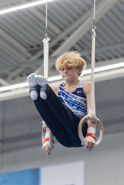 Young gymnast with blonde hair performs L-sit on rings, displaying focus and strength during training in indoor facility