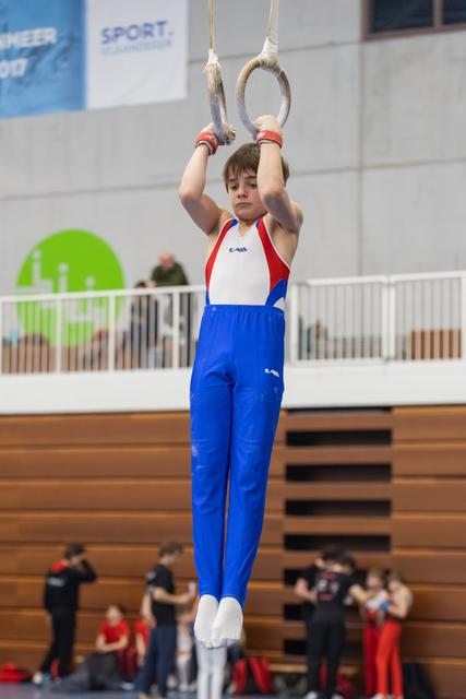 Young gymnast in red, white and blue uniform performs still rings routine with focused determination in indoor gymnasium