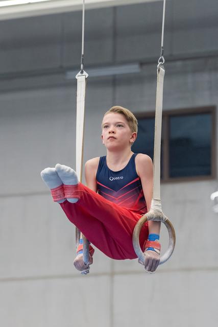Young gymnast performs rings routine with determined expression, wearing navy and red leotard in training facility