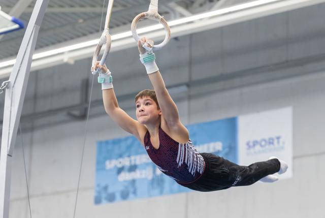 Young gymnast performing a horizontal hold on rings with focused expression, wearing purple leotard in indoor sports facility