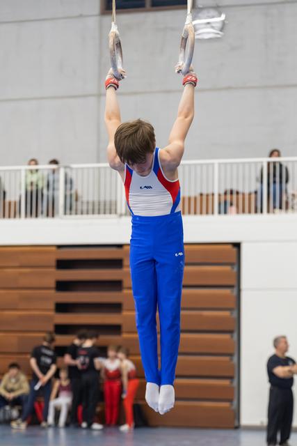 Young gymnast in blue and red uniform performs vertical hold on rings, demonstrating strength and focus in gymnasium
