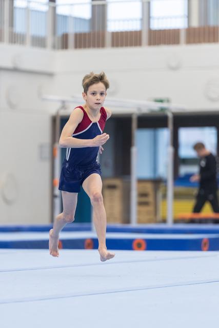 Young gymnast in navy leotard running barefoot across white floor mat during routine, focused expression, training facility