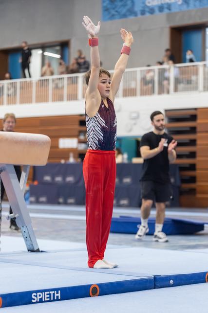 Young gymnast in red pants and patterned leotard stands at attention with arms raised, preparing for balance beam routine