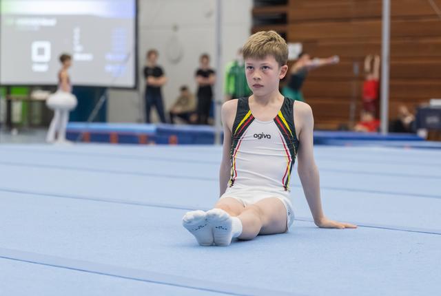 Young gymnast in white and black leotard sits on blue mat with focused expression, others training in background