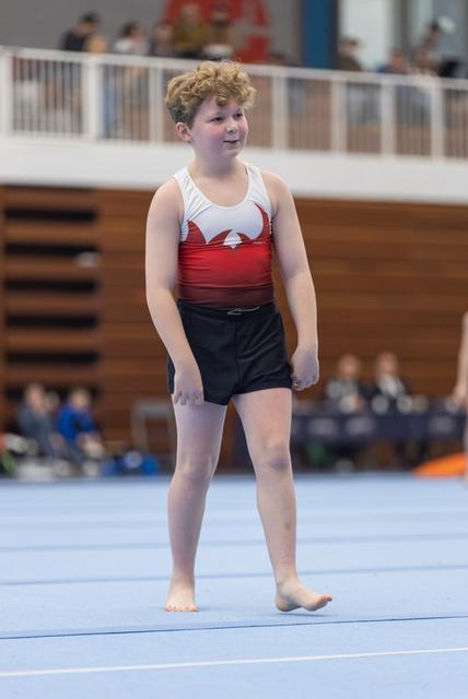 Young gymnast in red and white leotard smiles confidently while standing on floor exercise mat in gymnasium