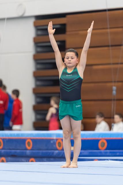 Young gymnast in teal and black leotard stands with arms raised overhead on floor exercise mat, showing focused expression