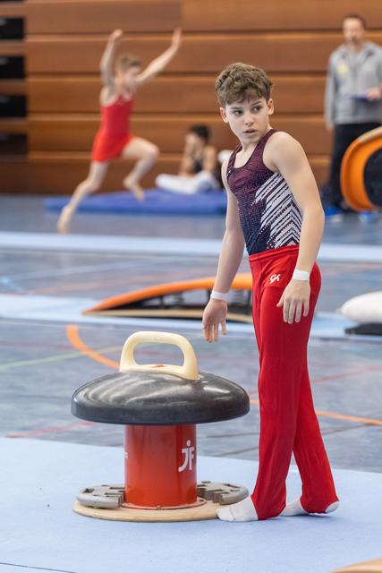 Young gymnast in red pants and navy leotard stands beside a mushroom trainer, focused and ready to practice