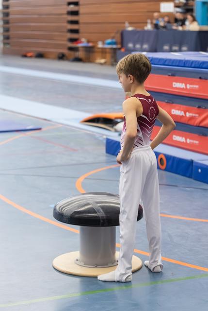 Young gymnast in burgundy leotard and white pants stands with hand on hip beside a mushroom trainer in the gym