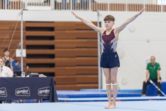 Young gymnast in maroon and navy leotard stands with arms raised in finishing pose on blue competition mat