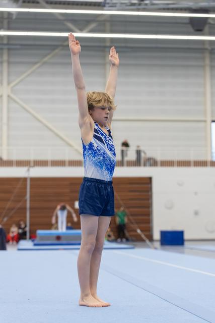 Young gymnast in blue and white leotard stands tall with arms raised overhead on floor mat in indoor gymnasium