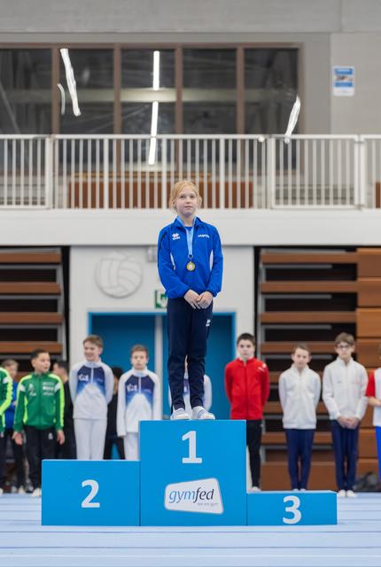 Young gymnast in blue tracksuit stands on first place podium wearing medal, with competitors lined up behind in gymnasium