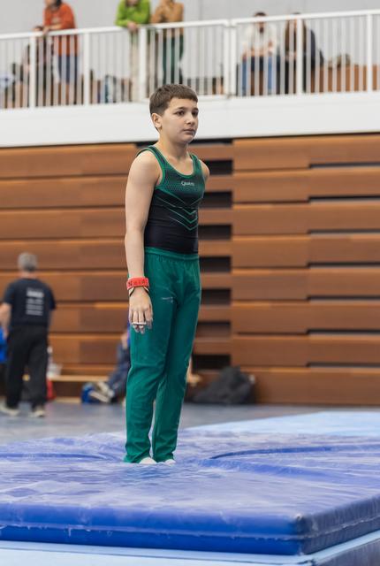 Young gymnast in teal uniform stands attentively on blue floor mat, waiting for his turn while spectators watch from bleachers