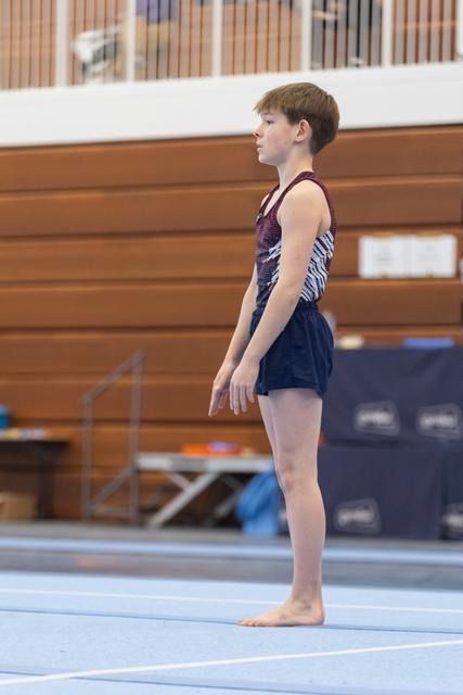 Young gymnast in blue leotard stands barefoot in ready position on floor mat, arms at sides, focused expression
