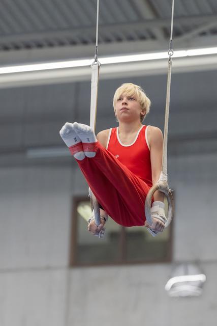 Young blonde gymnast performing on still rings in red uniform, displaying focused concentration and strength