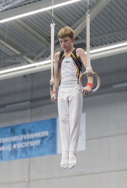 Young male gymnast performing on still rings, displaying intense concentration while holding position in white uniform