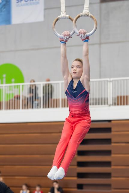 Young gymnast hangs from still rings during routine, displaying focused concentration and proper form in gymnastics facility