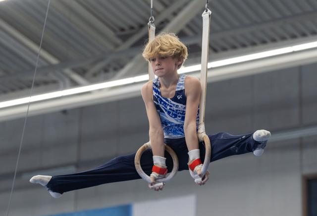 Young male gymnast performs on still rings with pointed toes and focused expression in indoor training facility