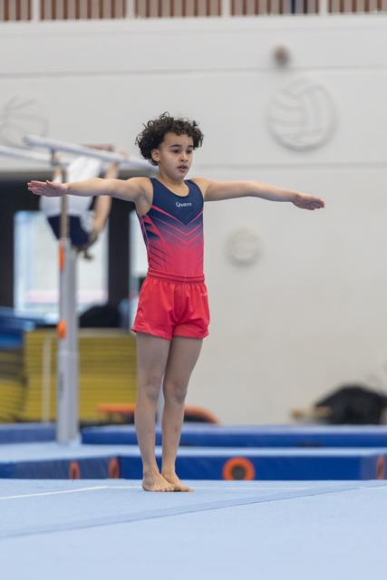 Young gymnast with curly hair performing a T-pose balance on floor mat, arms extended, displaying focused concentration