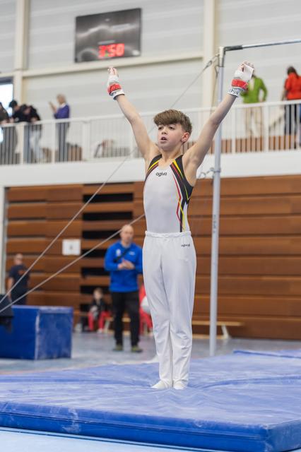 Young gymnast raises arms in triumph after completing high bar routine, wearing white and yellow striped leotard