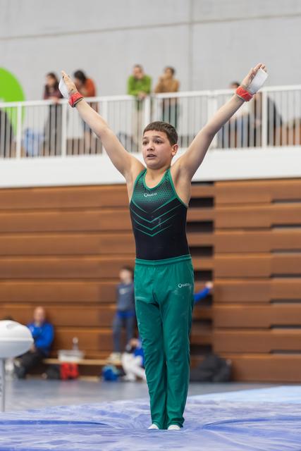 Young gymnast strikes a victorious finish pose with arms raised high, wearing teal and black leotard in gymnasium