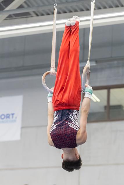 Young gymnast performing an inverted hang on still rings, wearing red top and patterned shorts in indoor training facility