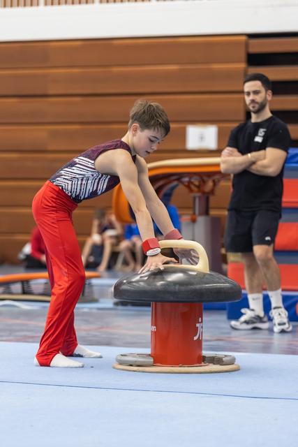 Young gymnast in red pants practicing vault technique on pommel horse while coach observes in gymnasium