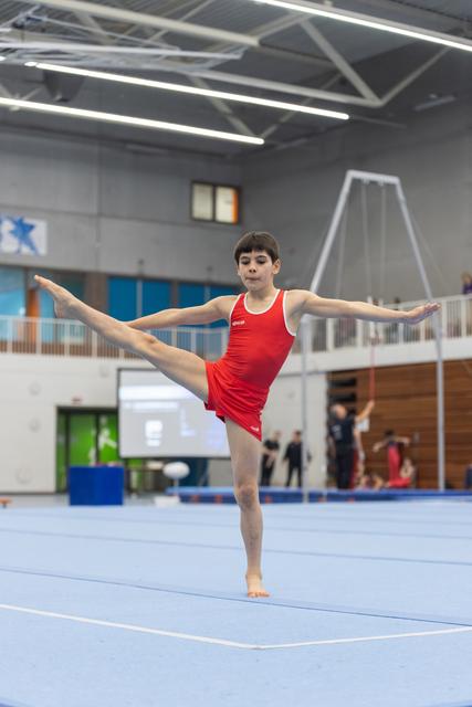 Young gymnast performs vertical split leap on floor exercise mat, arms extended in T-position at indoor training facility