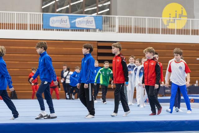 Young gymnasts in team tracksuits march in formation across a blue floor mat during opening ceremony in a gymnasium