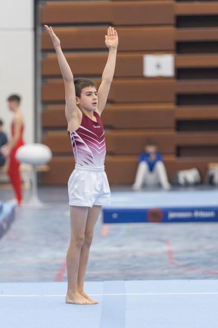Young gymnast in maroon leotard stands with arms raised overhead on floor mat, displaying focused expression during routine
