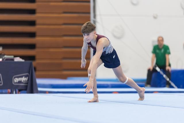 Young male gymnast performing floor routine, landing from jump with focused expression and bent knees on blue mat