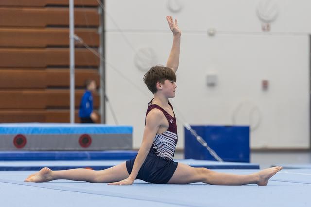 Young male gymnast performs a split on floor exercise mat with one arm raised, demonstrating flexibility in training facility